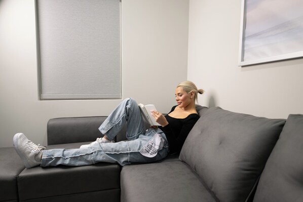 A person sitting on a grey sofa in a room with a plain wall and a window.
