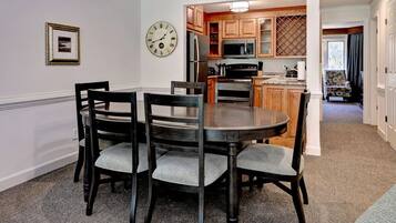 A dining area with a round table and four chairs, a clock on the wall, and a kitchen in the background.