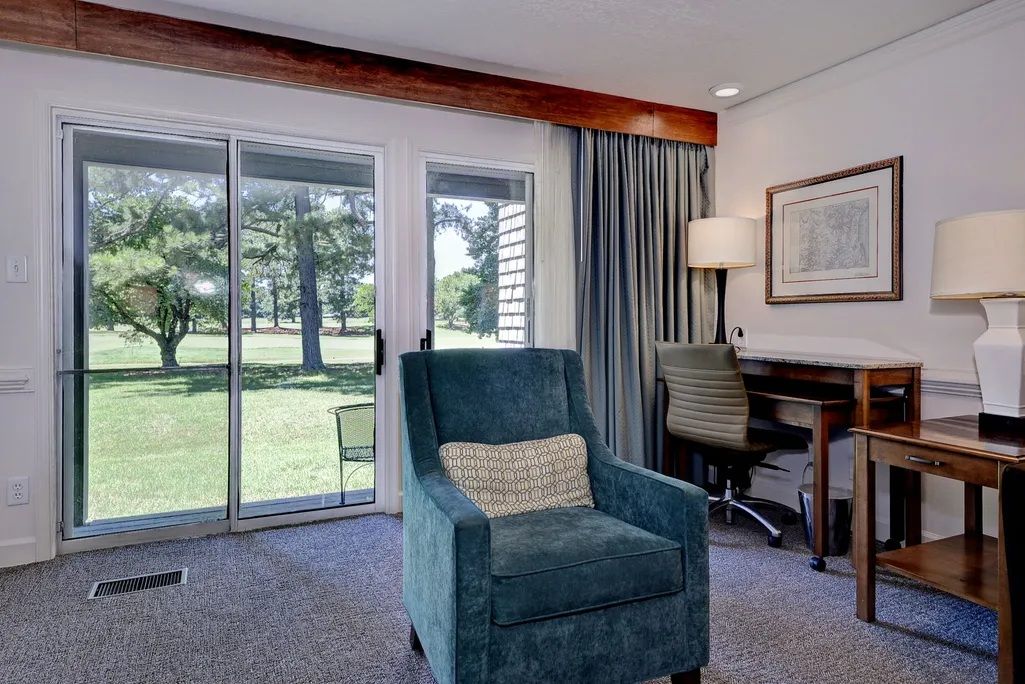 A room with a teal armchair, a desk, a chair, and a large window with a view of a lawn and trees.