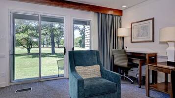 A room with a teal armchair, a desk, a chair, and a large window with a view of a lawn and trees.