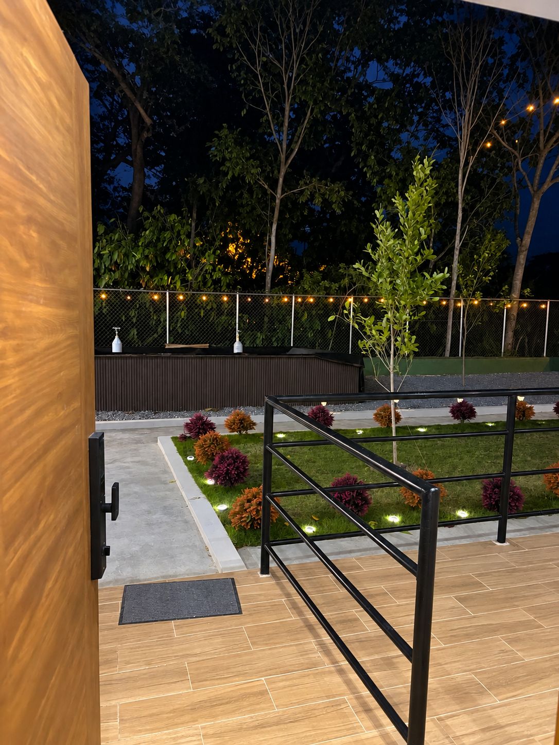 Eine Holztür mit schwarzem Griff, ein Metallzaun, ein gepflasterter Weg und ein Garten mit Zierpflanzen und Lichterketten.