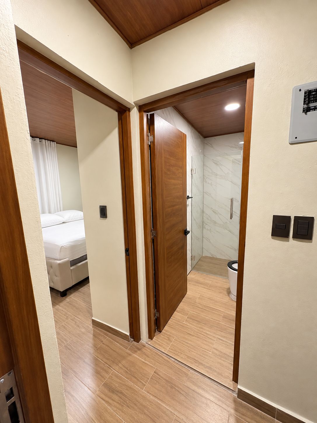 A hotel room with a wooden floor, a bed, a bathroom with a toilet, and a wall-mounted air conditioning unit.