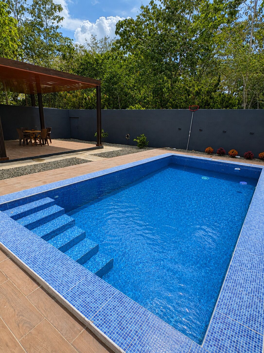 A rectangular swimming pool with blue mosaic tiles, surrounded by a patio area with a wooden pergola and a table with chairs.