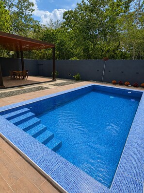 A rectangular swimming pool with blue mosaic tiles, surrounded by a patio area with a wooden pergola and a table with chairs.