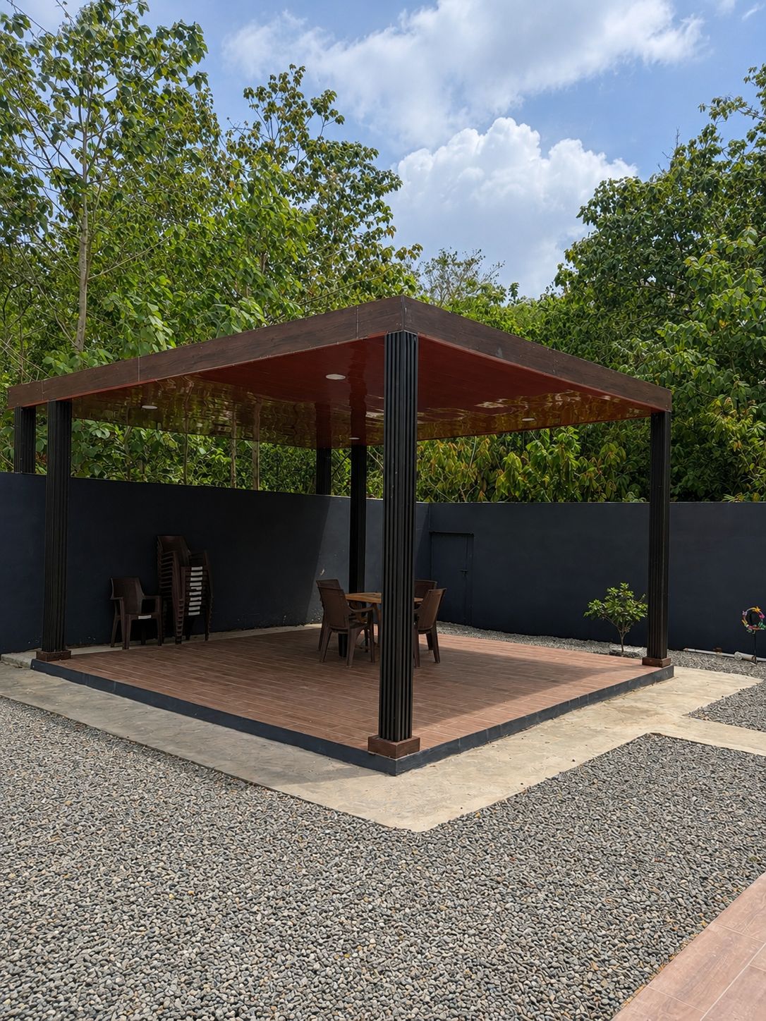 Un área de estar al aire libre con techo, con una mesa y sillas, rodeada por un muro bajo y un suelo de grava.
