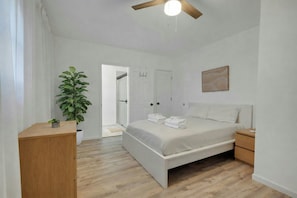A modern bedroom with a wooden floor, a bed with white linens, a bedside table, a nightstand, a ceiling fan, and a large potted plant. - Dog Friendly Lakeview Family Retreat with Hot Tub, Firepit and Game Room (Jefferson)