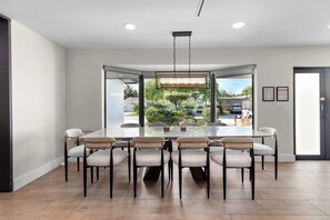 A modern dining area with a glass table surrounded by chairs, a hanging light fixture, and a view of the outdoors. - Waterfront Pool I Paddleboards, Firepit + Fishing (Miami)