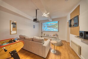 A modern living room with a sofa, a bean bag, a projector screen displaying a scenic image, a dartboard, and a small table with decorative items.