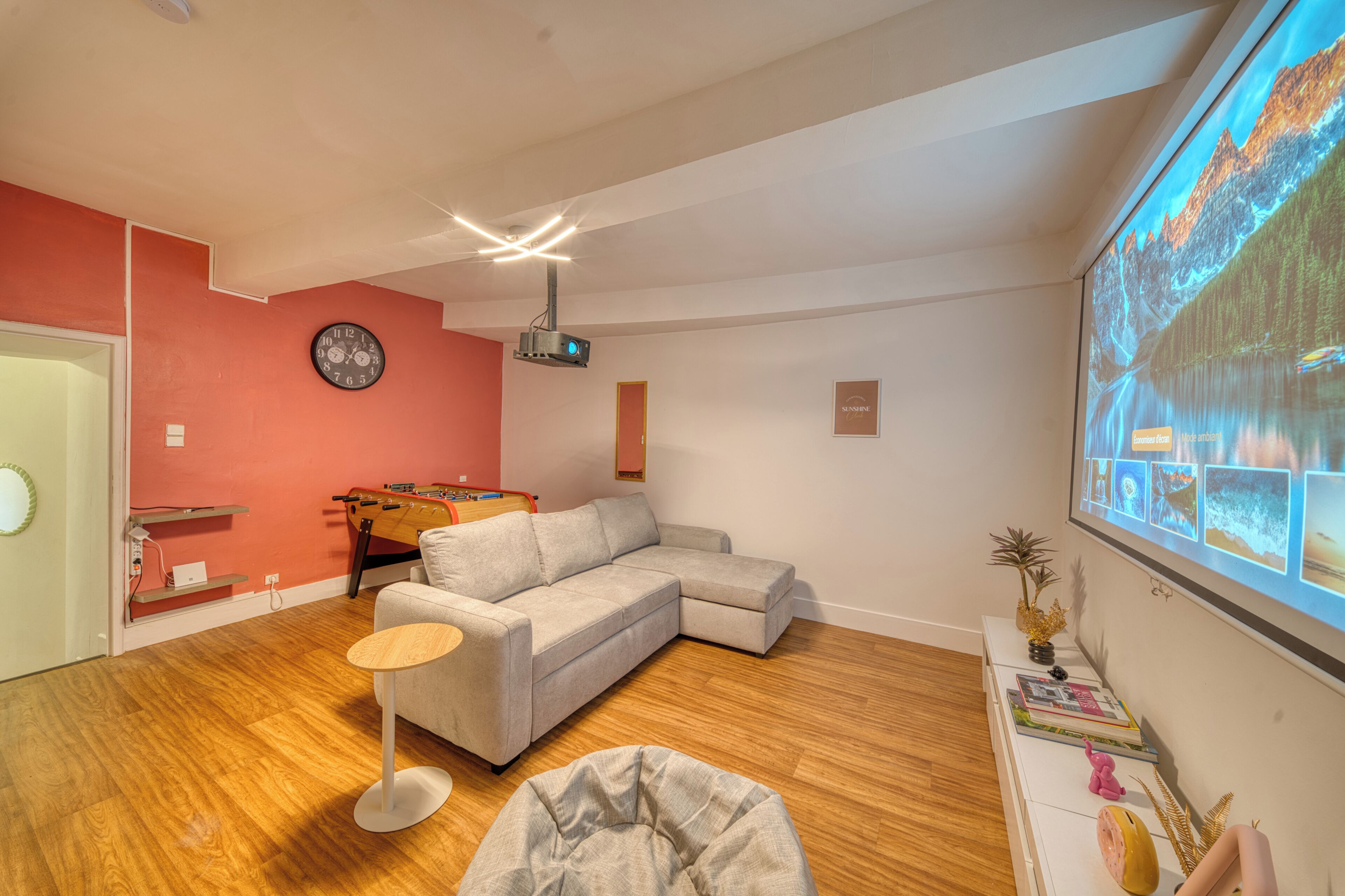 Een moderne woonkamer met een hoekbank, een tafelvoetbaltafel, een projectorscherm waarop een mooi landschap te zien is, en een wandklok.