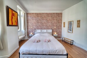 A bedroom with a brick-patterned wall, a bed with white bedding, two bedside tables with lamps, a bench, and a painting on the wall.