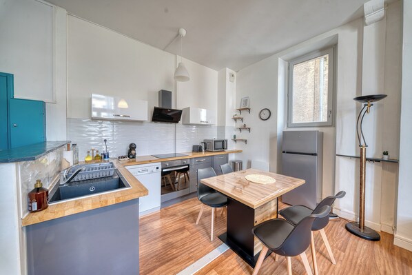 A compact kitchen with a dishwasher, microwave, and a refrigerator. There is a dining table with chairs and a small window providing natural light.