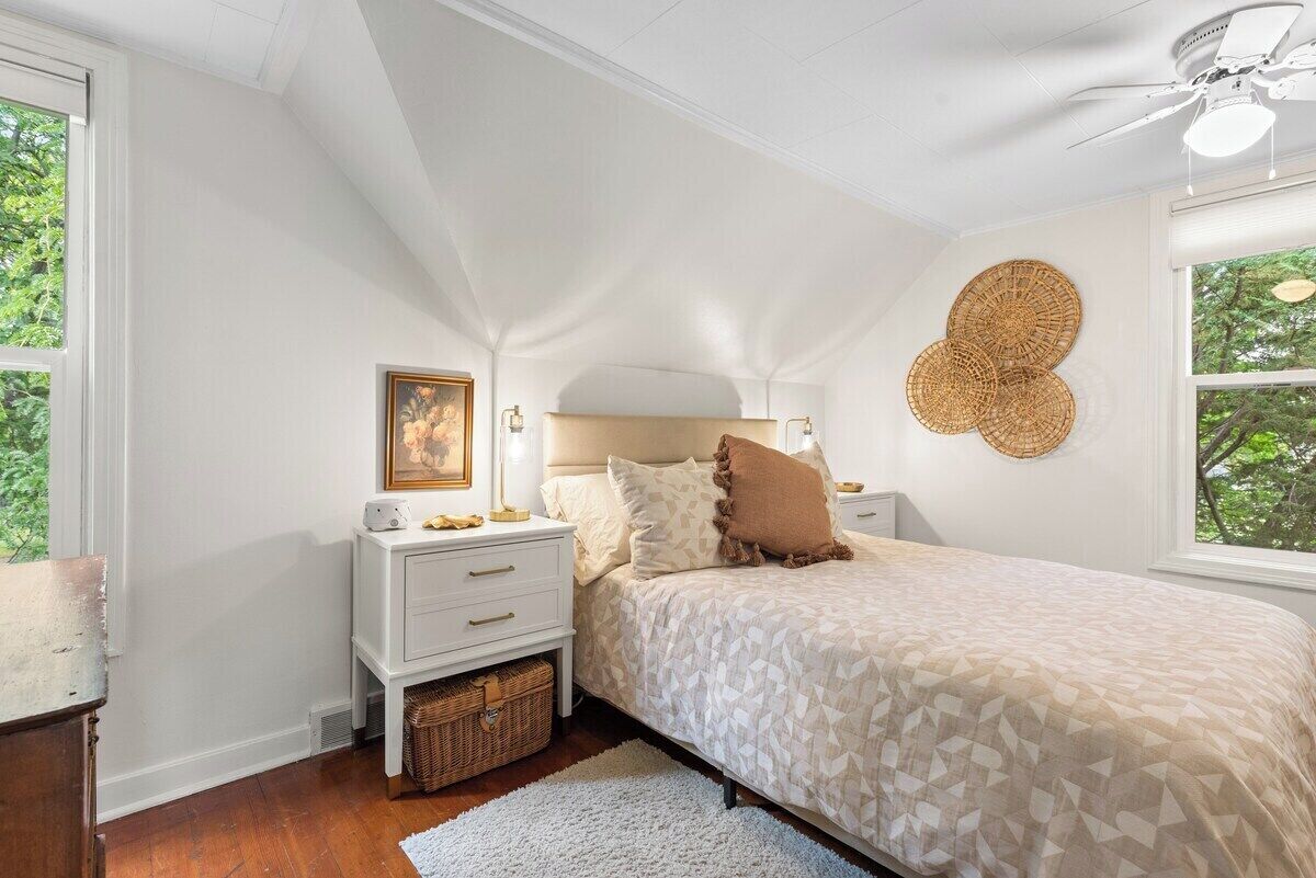 A bedroom with a bed, bedside tables, a nightstand, a ceiling fan, and a window with a view of trees.