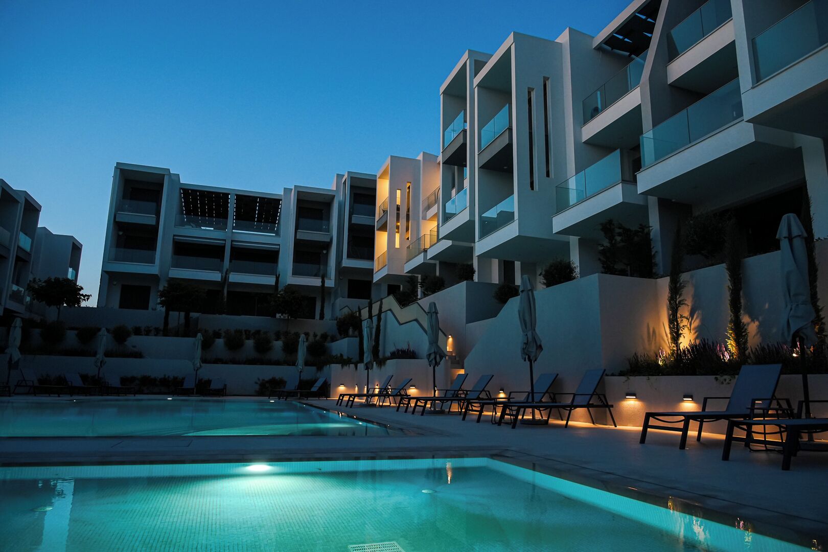 A modern hotel with a swimming pool, lounge chairs, and well-lit balconies.
