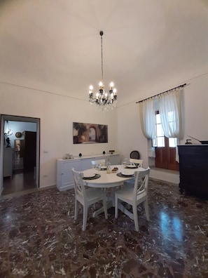 A dining area with a round table, white chairs, a chandelier, and a painting on the wall. - Adorable 2-bedroom apartment with AC in the center of charming Noto (Noto)