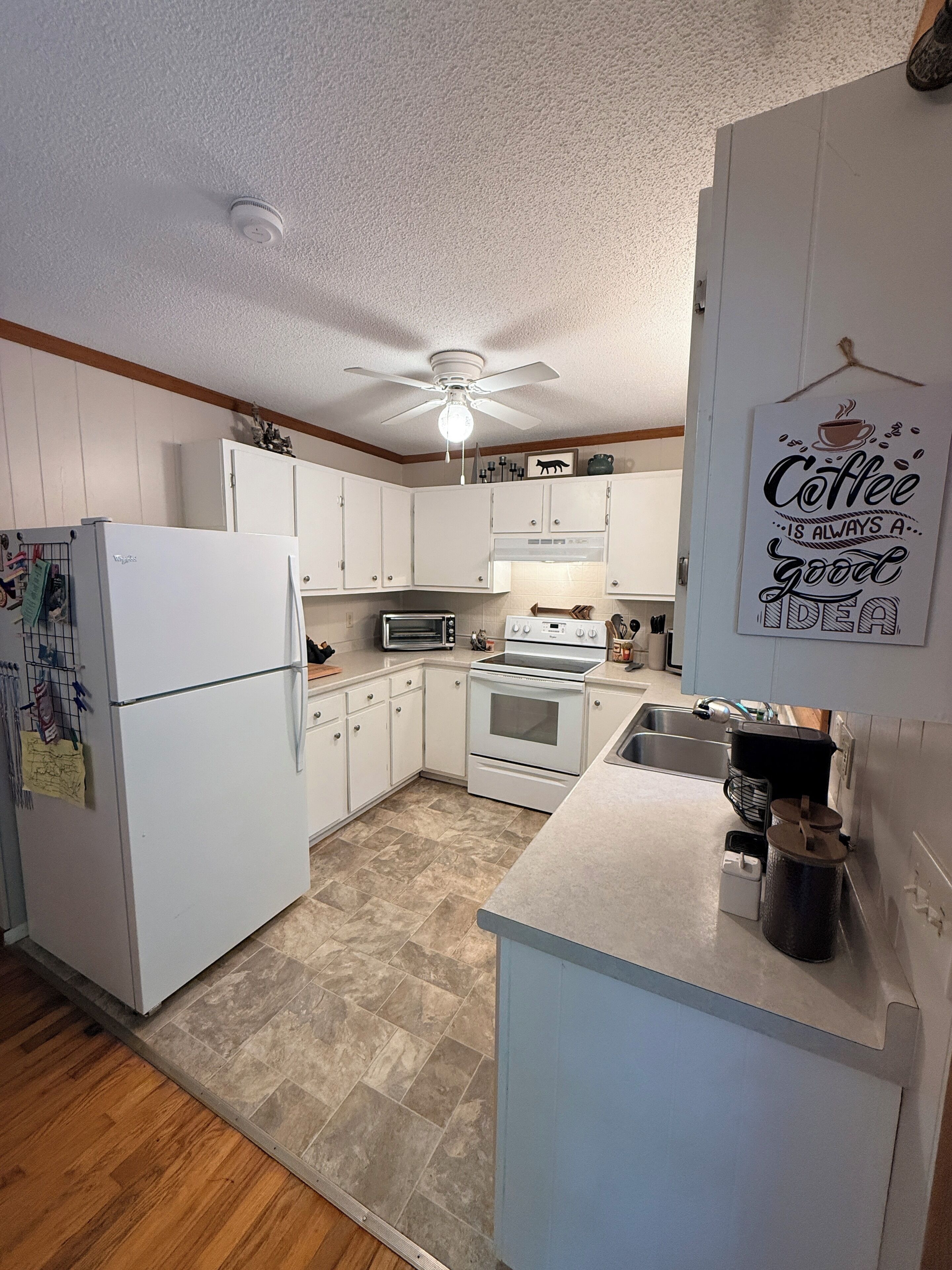 Une cuisine avec des meubles blancs, un réfrigérateur, un four à micro-ondes, un four traditionnel, un ventilateur de plafond et une décoration murale sur le thème du café.