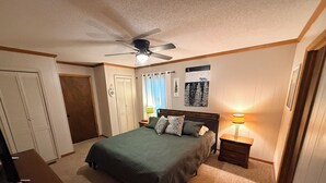 Quarto com cama, mesa de cabeceira, ventilador de teto, abajur e porta.