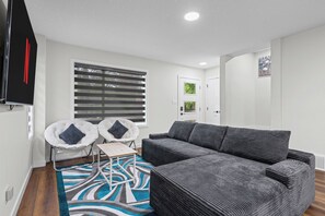 Ein modernes Wohnzimmer mit einem grauen Ecksofa, zwei weißen Sesseln mit schwarzen Kissen, einem kleinen Holztisch und einem gemusterten Teppich.