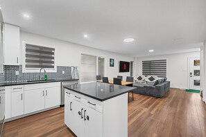A modern kitchen with white cabinets, a black countertop, and a dining area with a grey sofa and wooden flooring. - 1BHK Close to Downtown/Whyte Ave/UOFA 10922 (Edmonton)