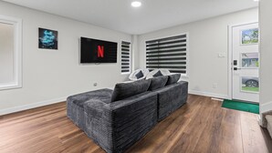 Un salon moderne avec un canapé gris, un téléviseur à écran plat et une grande fenêtre équipée de stores.