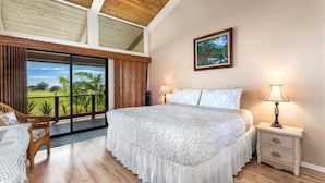 A bedroom with a large bed, a nightstand, a lamp, a painting, and a view of a green landscape through a sliding door. - Waikoloa Villas#a203:top Floor Beauty, Ocean Views (Waikoloa)