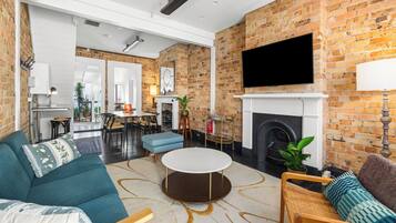 A modern living room with a brick wall, a fireplace, a flat-screen TV, and a dining area.