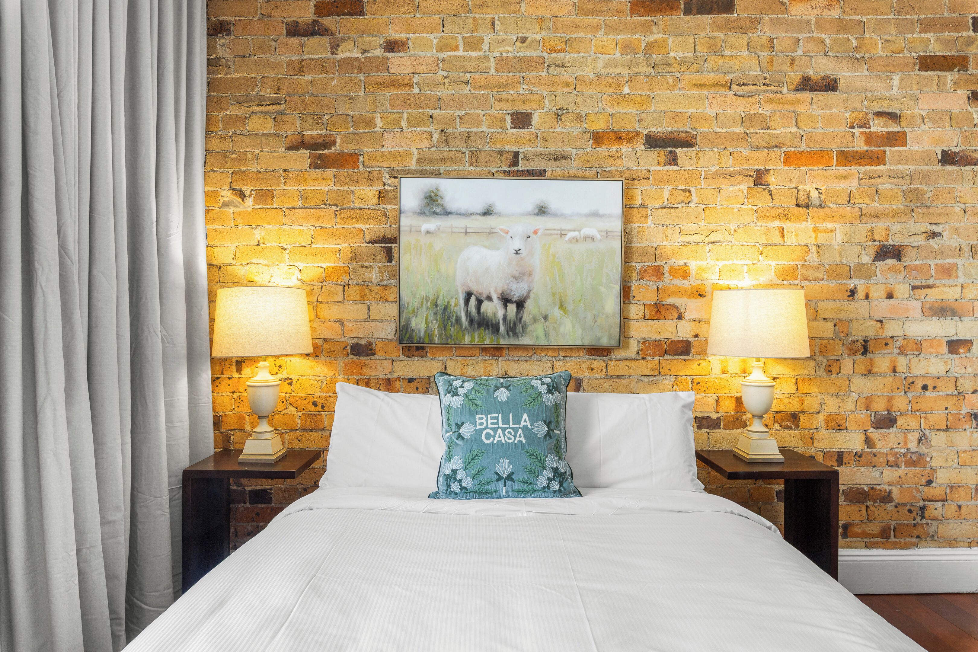 A hotel room with a brick wall, a framed pastoral painting, two bedside lamps, a white bedspread, and a pillow with 'Bella Casa' text.