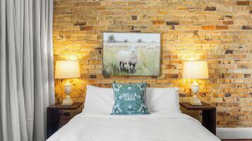 A hotel room with a brick wall, a framed pastoral painting, two bedside lamps, a white bedspread, and a pillow with 'Bella Casa' text.