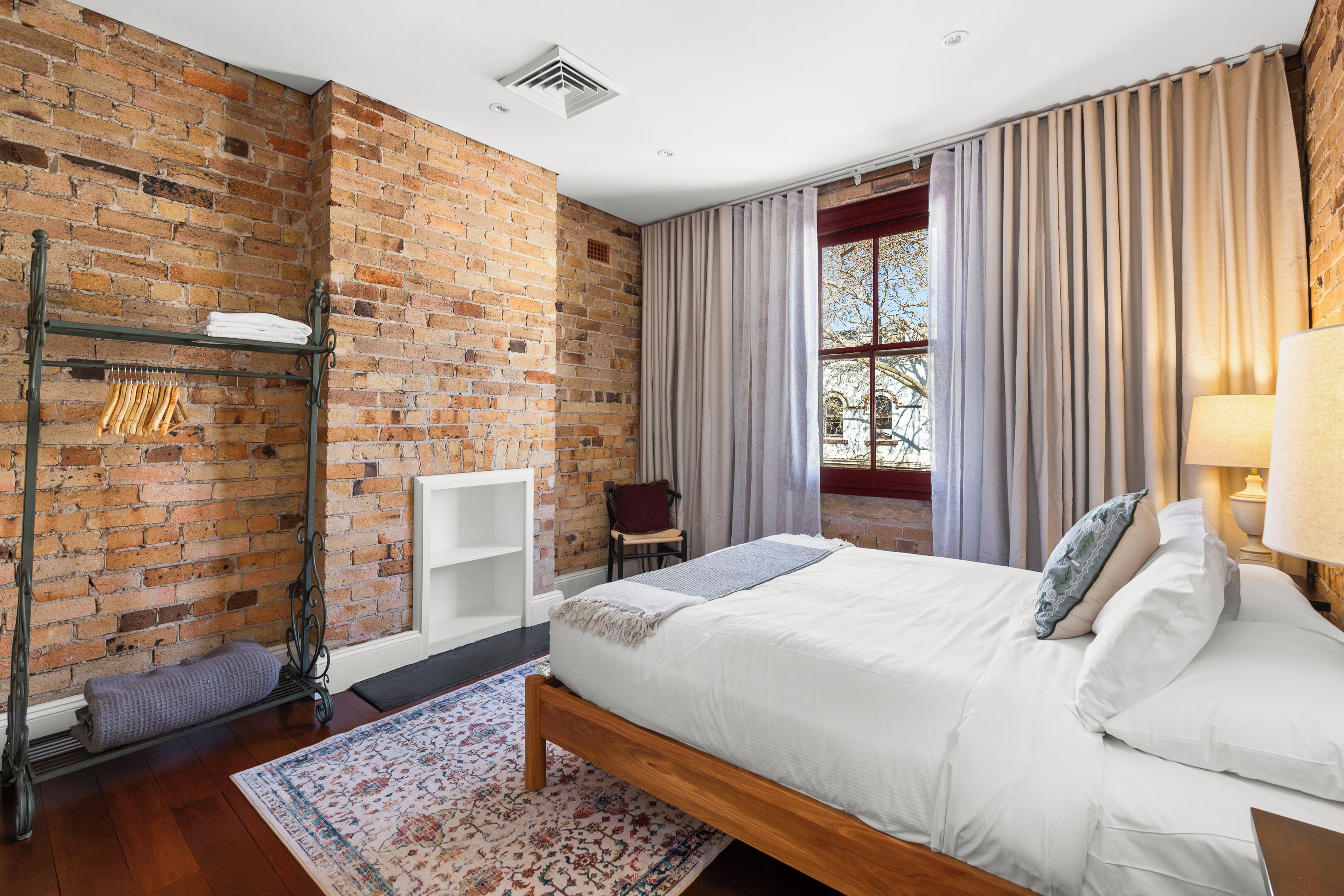 Une chambre avec un mur en briques, un lit avec une literie blanche, une tête de lit en bois, une fenêtre avec des rideaux et un radiateur.