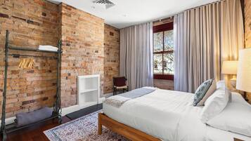 A bedroom with a brick wall, a bed with white bedding, a wooden headboard, a window with curtains, and a radiator.