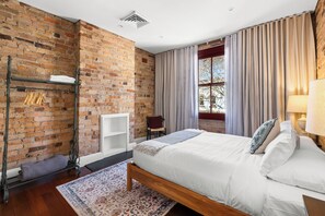 A bedroom with a brick wall, a bed with white bedding, a wooden headboard, a window with curtains, and a radiator.