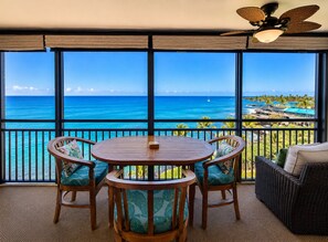 A balcony with a view of the ocean and a resort. There is a round wooden table with chairs and a wicker sofa. - Kona Alii#704: 7th Floor Penthouse, Oceanfront! (Kailua-Kona)