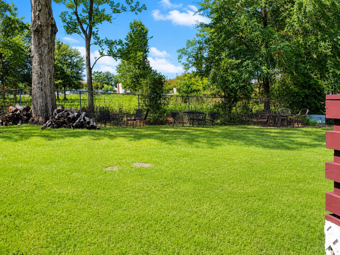 A grassy outdoor area with a picnic table and chairs, surrounded by trees and a fence.