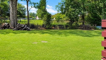 A grassy outdoor area with a picnic table and chairs, surrounded by trees and a fence.
