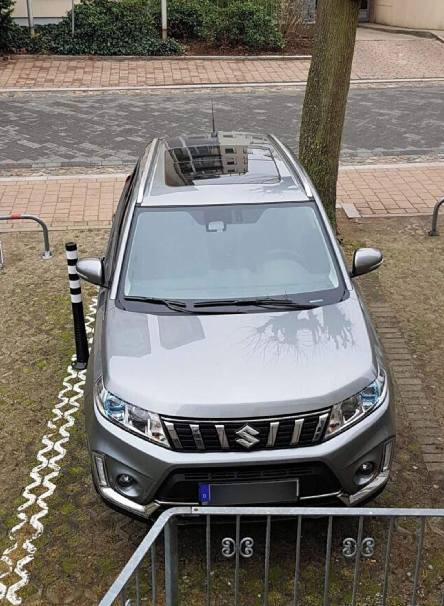 Ein silberfarbenes Suzuki-Auto steht auf einem befestigten Untergrund. Im Vordergrund sind ein Baum und ein Metallzaun zu sehen.