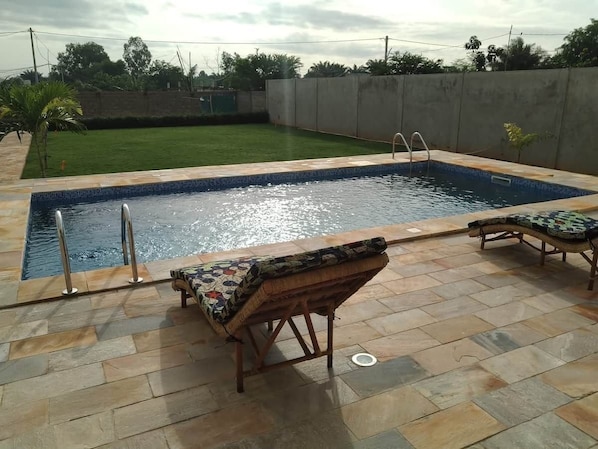 A swimming pool with a tiled deck, two lounge chairs, and a concrete wall in the background.