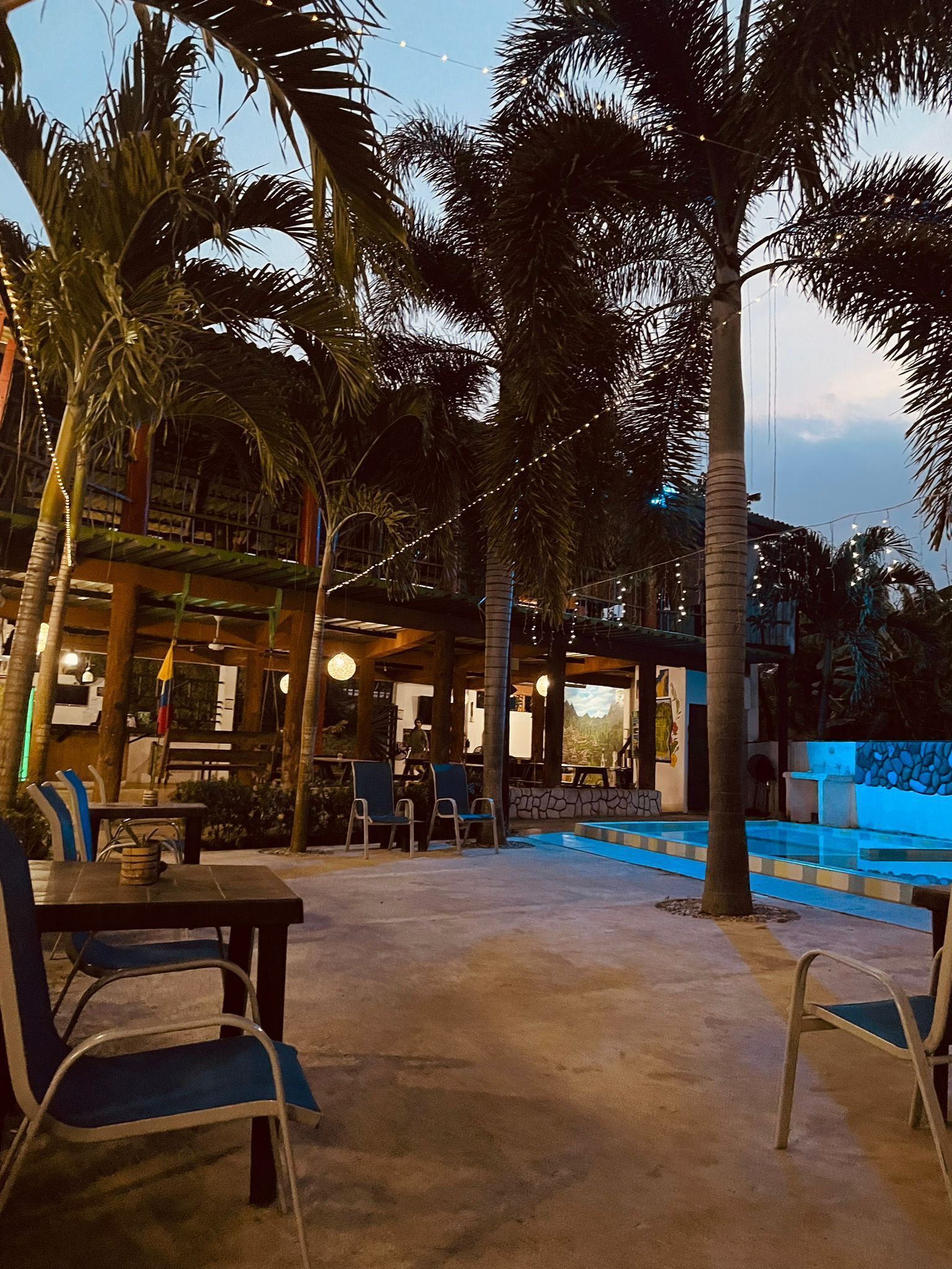 Un espacio al aire libre con mesas, sillas, piscina y palmeras.