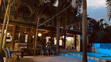 Un espacio al aire libre con mesas, sillas, piscina y palmeras.