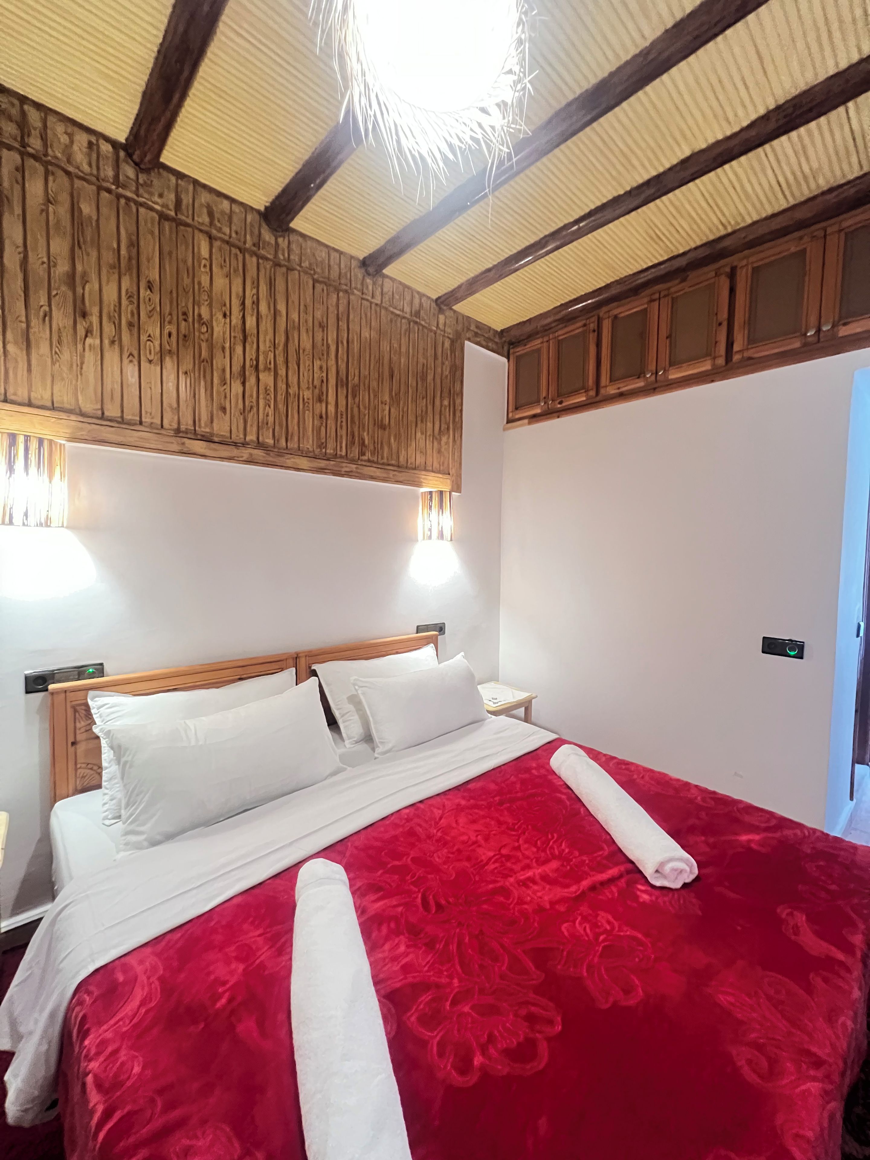 A neatly made bed with white linens and a red bedspread, wooden headboard, and a ceiling with exposed beams.