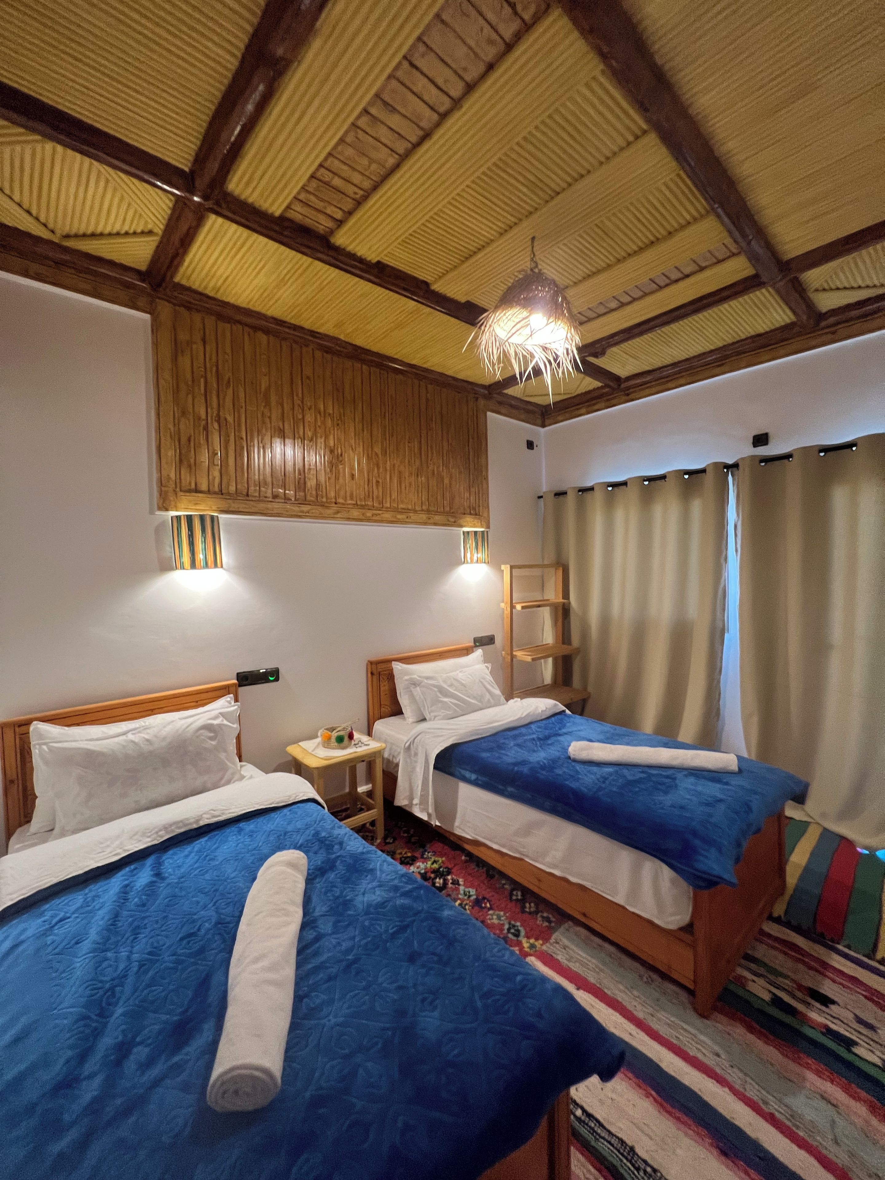 A room with two beds, wooden headboards, a ceiling with wooden beams, a hanging light fixture, a small bedside table with a lamp, and a colorful rug.