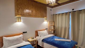 A room with two beds, wooden headboards, a ceiling with wooden beams, a hanging light fixture, a small bedside table with a lamp, and a colorful rug.