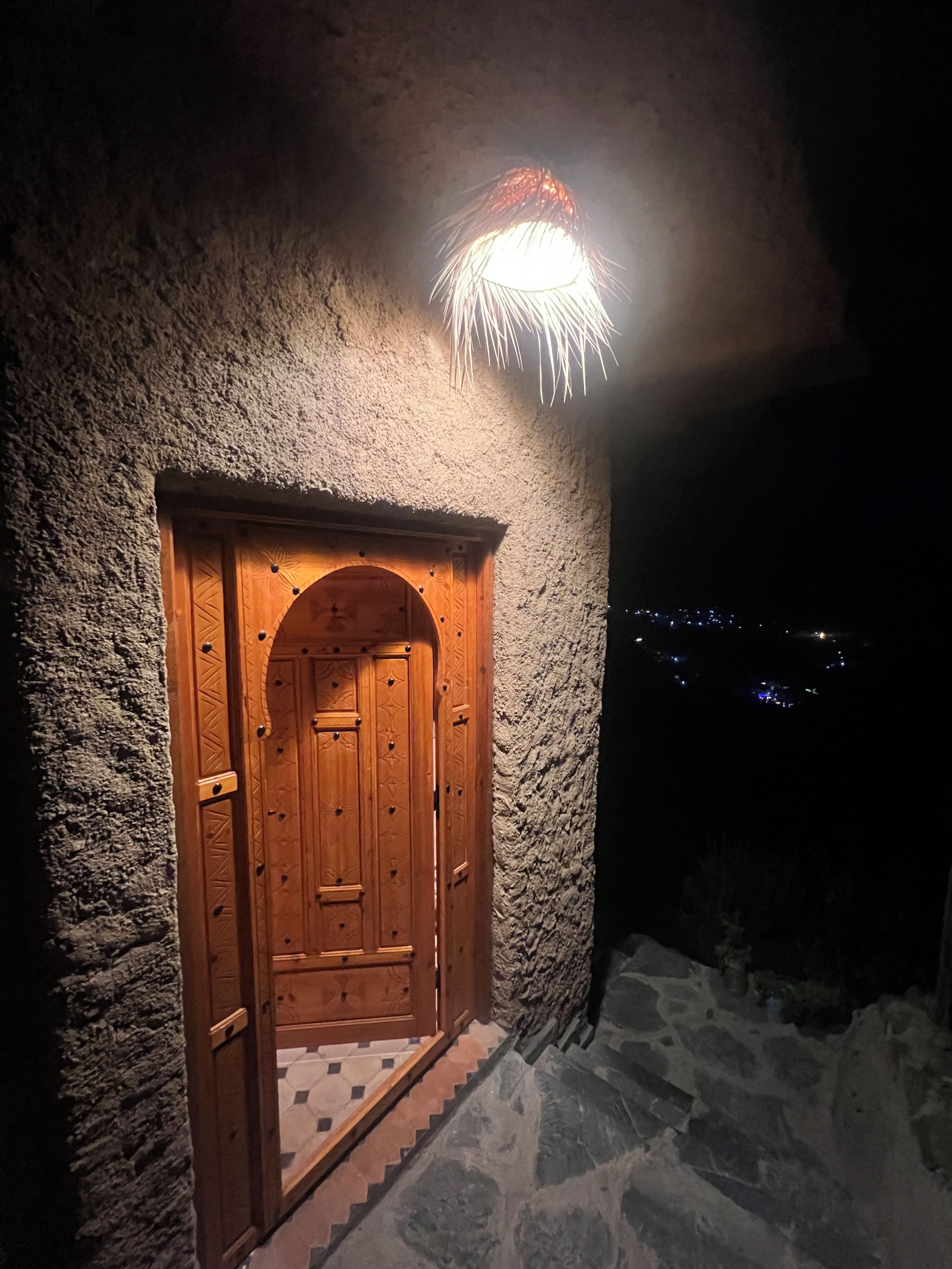 Une porte en bois surmontée d’une arche décorative, intégrée dans un mur de pierres à la texture particulière, et qui donne sur un escalier.