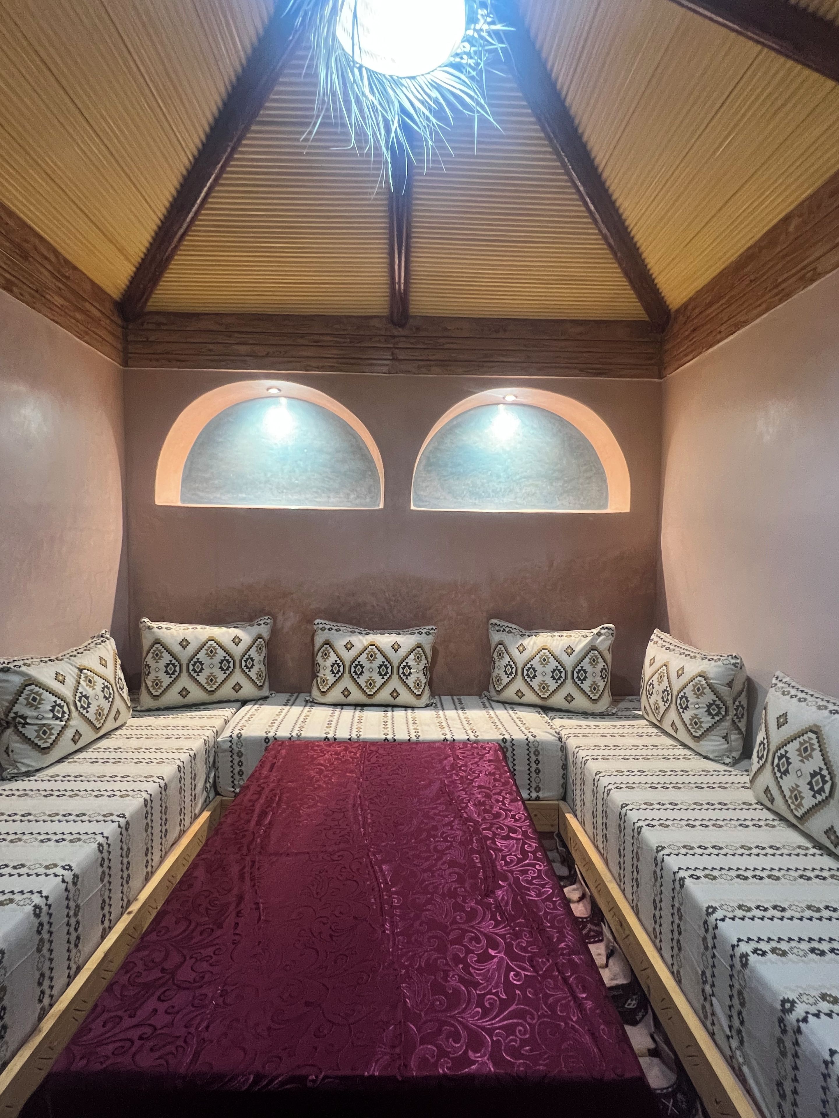 Una habitación con techo alto, dos ventanas arqueadas, un sofá largo con cojines estampados y una manta roja.