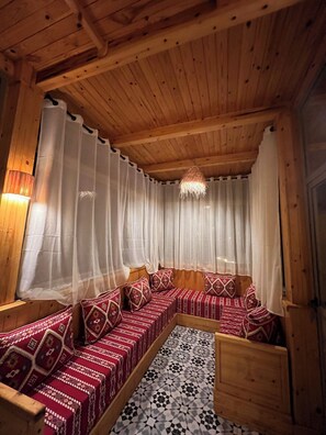 A wooden cabin interior with a long, narrow sofa set against a wall, a patterned floor, and a wooden ceiling.