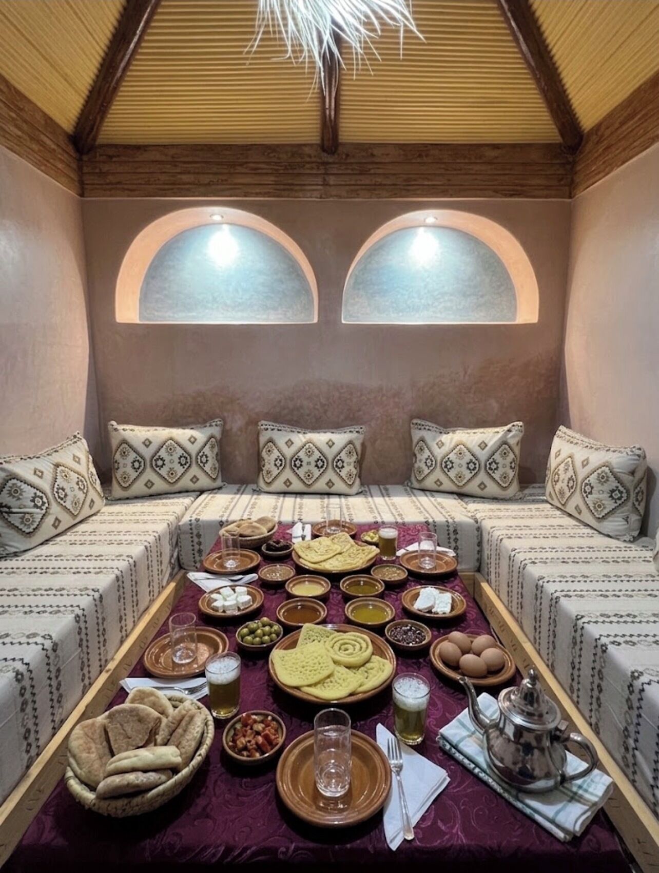 Un coin salon accueillant, avec un plafond bas, deux fenêtres en arc de cercle et une table dressée avec différents plats et boissons.