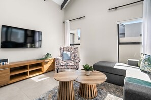 A modern living room with a flat-screen TV, a floral armchair, a grey sofa, and a wooden coffee table.