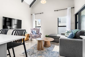 A modern living room with a flat-screen TV, a dining table with chairs, a sofa, a patterned armchair, and a coffee table.