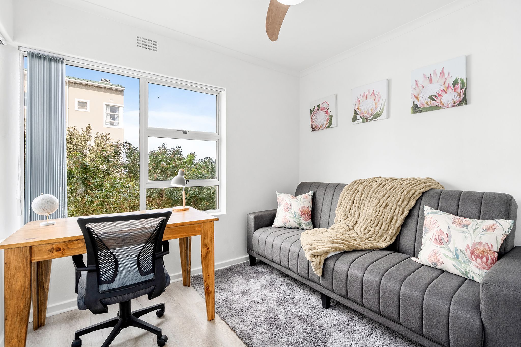 A modern living room with a grey sofa, a wooden desk, and a black office chair. There are floral wall art pieces and a view of greenery outside.