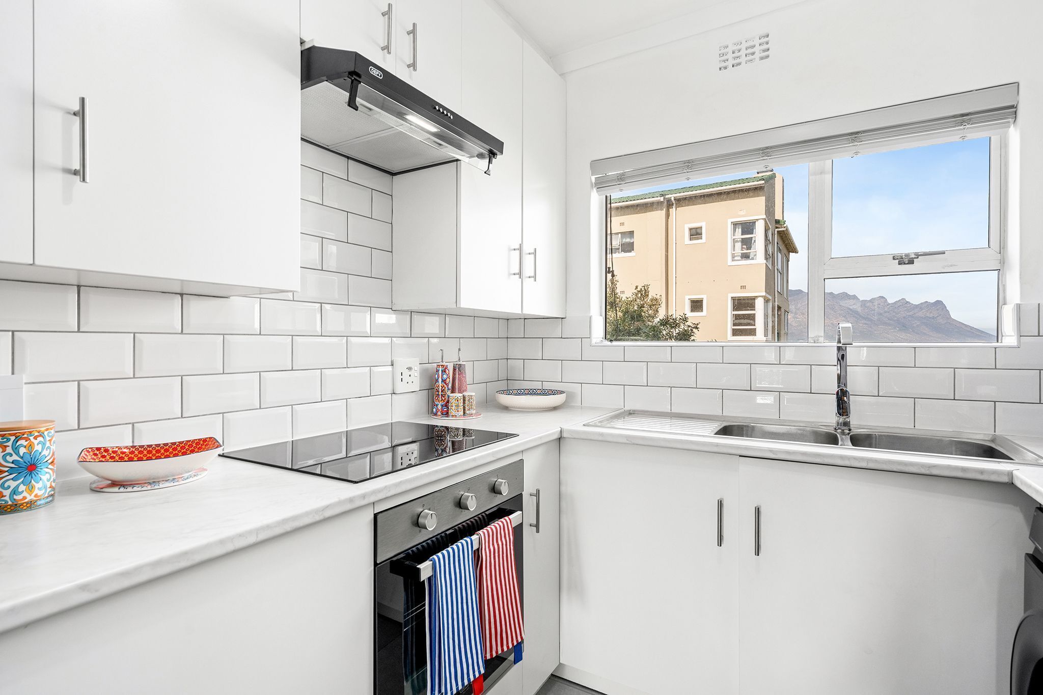 Una cuina moderna amb armaris blancs, forn integrat, fogó i finestra amb vista a un edifici i muntanyes.