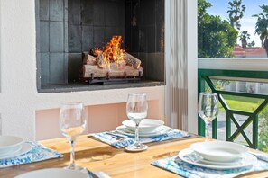 Un área de comedor con una mesa lista para dos personas, una chimenea con leña encendida y vista a palmeras a través de una ventana.