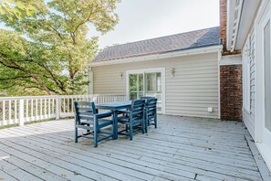 A covered outdoor deck with a table and chairs, a white fence, and a building with a brick section.